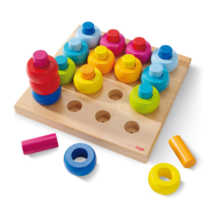 The Rainbow Whirls Wooden Sorting and Stacking Game by HABA features a square base with pegs and colorful rings in red, pink, blue, yellow, orange, and green. Ideal for early childhood development, some rings are stacked while others scatter—a delightful Montessori toy for learning and play.