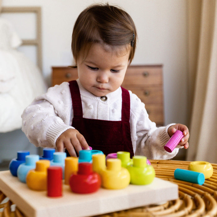 A toddler in a white sweater and red overalls plays with the colorful Rainbow Whirls Wooden Sorting and Stacking Game by HABA on the table. The room, featuring a wooden dresser and beige curtains, creates a cozy atmosphere as the child joyfully works on fine motor skills with a cylindrical block.