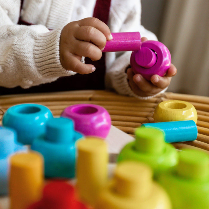 A child in a white sweater plays with HABAs Rainbow Whirls Wooden Sorting and Stacking Game on a table, enhancing fine motor skills with its bright blue, pink, yellow, and orange pieces as the child carefully assembles a pink piece into another structure.