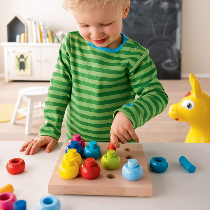 A child in a green striped shirt plays with the HABA Rainbow Whirls Wooden Sorting and Stacking Game on a board. A yellow toy horse is in the background, near shelves filled with books and toys. The child smiles, focused on arranging the pieces for fine motor development.