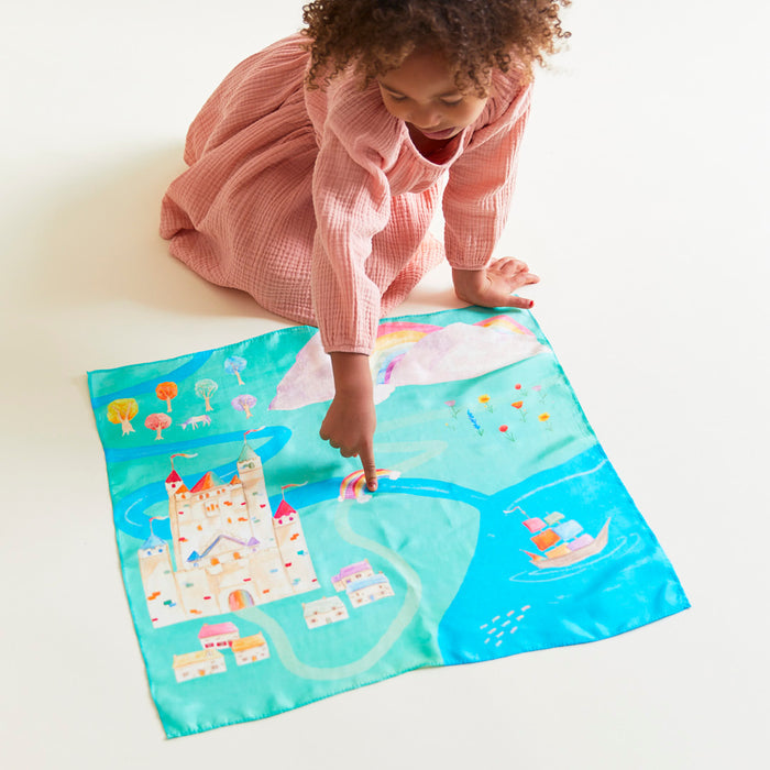 A child in a pink dress kneels on a white surface, pointing at Sarahs Silks Rainbowland Play Map, featuring a castle, ship, rainbow, trees, and houses. The vibrant scene is tailor-made for imaginative play akin to a storybook illustration.