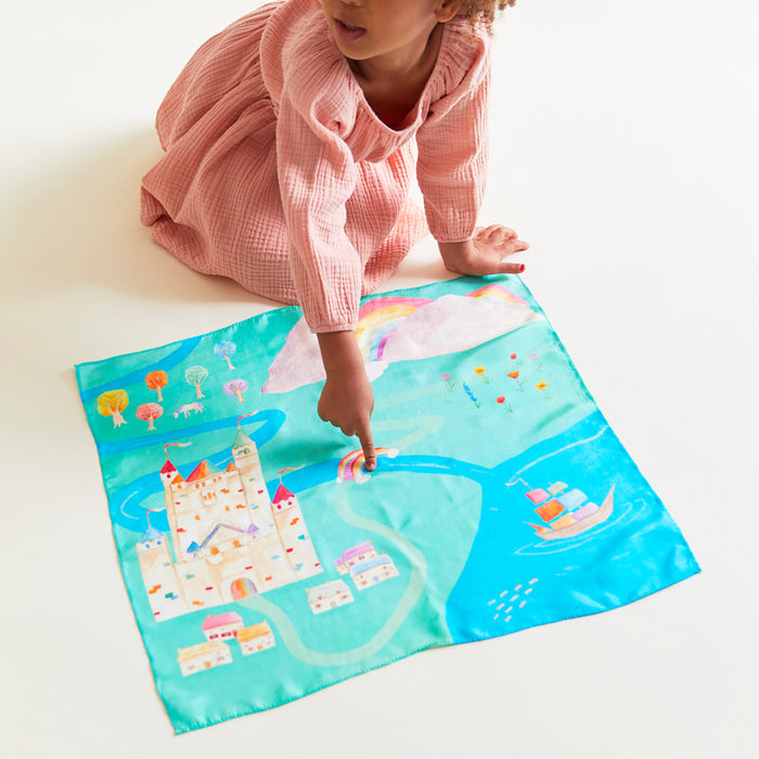 A child in a pink dress sits on the floor pointing at Sarahs Silks Rainbowland Play Map, a colorful mat featuring a castle, trees, river, and sailboat on a vibrant blue background. Playsilks add more adventure to this imaginative play scene.