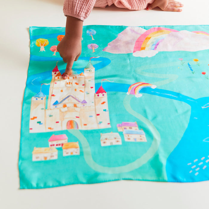 A childs hand points to a colorful illustration on fabric, featuring a castle, rainbow, and trees against a turquoise backdrop. This Rainbowland Play Map by Sarahs Silks resembles a whimsical map, sparking imaginative play with its vibrant landmarks.