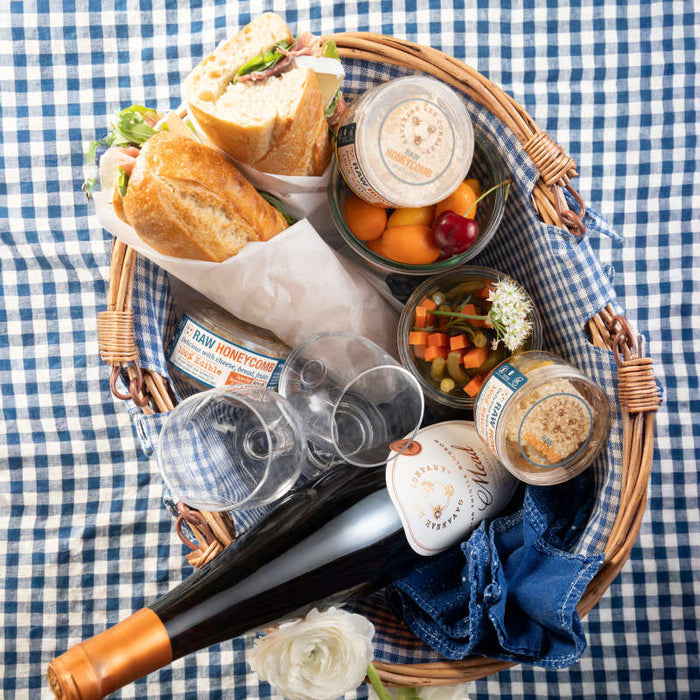 A Savannah Bee Companys Raw Honeycomb Picnic Round sits on a blue checkered cloth, containing a sliced sandwich, jars with spreads and honeycomb, assorted fruits, two empty glasses, a bottle of wine, and a white flower for decoration.