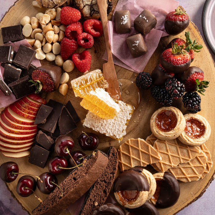 The Savannah Bee Companys Raw Honeycomb Picnic Round is a rustic wooden platter ideal for picnics, featuring assorted chocolates, honeycomb, berries, cherries, apple slices, nuts, cookies, wafers, dark chocolate pieces and chocolate-covered strawberries for an energizing snack.