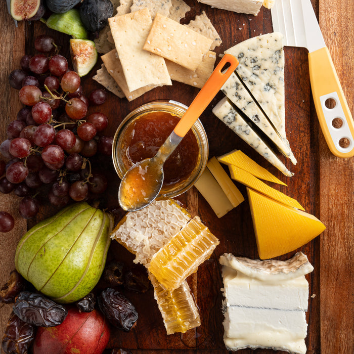 The Savannah Bee Companys Raw Honeycomb Picnic Round serves up a wooden board with cheeses, crackers, red grapes, sliced pear, pomegranate, honeycomb, figs, dates, a jam jar with a spoon, and includes a cheese knife on the side.