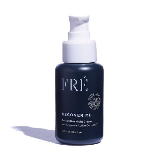 A dark blue bottle of FRÉ Skincares RECOVER ME Restorative Night Cream with a white pump top sits elegantly. The label showcases the Argania Active Complex™ for optimal skin repair, and its subtle shadow on a plain white backdrop enhances its sophistication.
