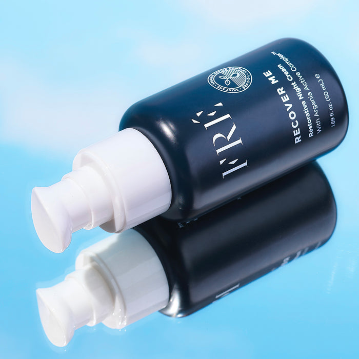 A dark blue FRÉ Skincare RECOVER ME Restorative Night Cream bottle with a white pump lies on a reflective blue surface, displaying the product and its clear reflection.