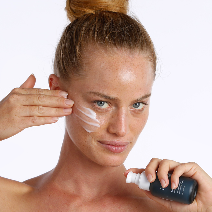 A woman with light skin and blue eyes smiles as she applies FRÉ Skincare’s RECOVER ME Restorative Night Cream to her cheek, holding the pump bottle in one hand against a plain white background.
