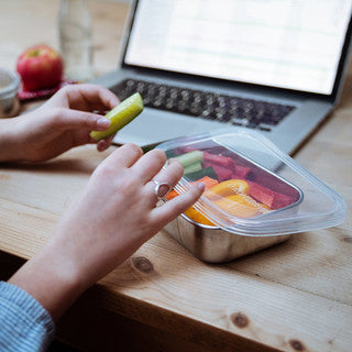 A person sits at a wooden table with an open laptop, holding a cucumber slice beside a U-Konserve Rectangle Container filled with assorted vegetables, an apple, and a small jar for plastic-free food storage.