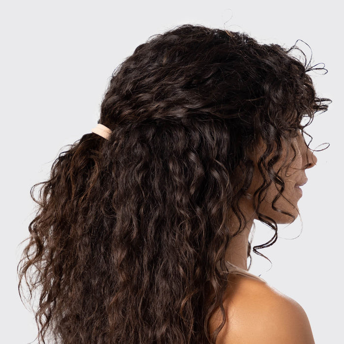 A person with long, curly brown hair is shown from the side, wearing a light pink Kitsch Recycled Nylon Hair Elastic. This eco-friendly accessory offers style and sustainability against a plain gray background.