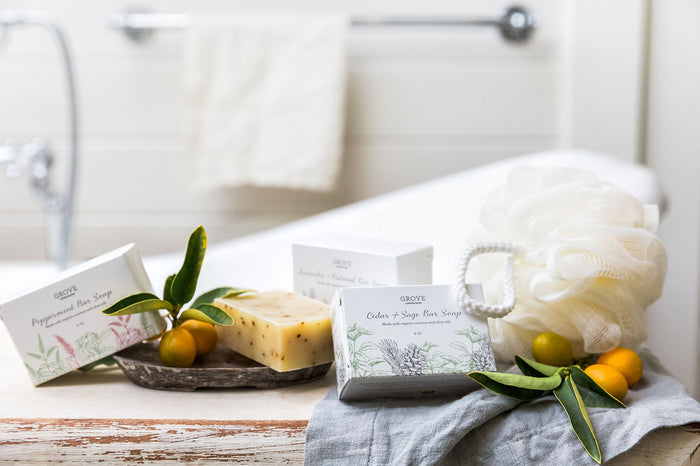 A rustic bathroom scene highlights Grove Co. soap bars in cardboard boxes on wood, with luxurious suds. A natural soap bar sits on a leaf-adorned tray next to a Grove Co. Recycled Plastic Loofah. Fruits and greenery add freshness near the bathtub, enhancing the serene ambiance.