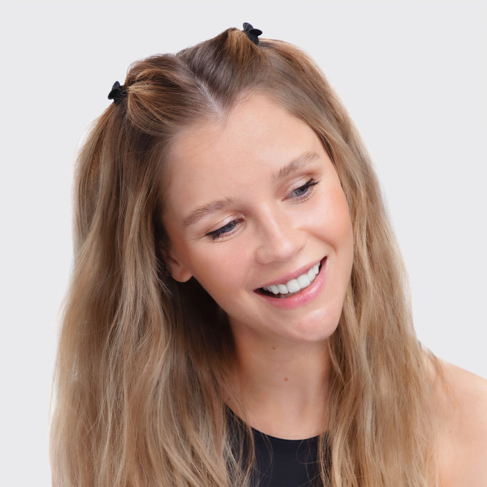 A smiling woman with long blonde hair styled in half-up, half-down pigtails wears chic Kitsch Recycled Plastic XS Claw Clips. Her sleeveless black top contrasts sharply against the plain white background as she gazes slightly to the side.