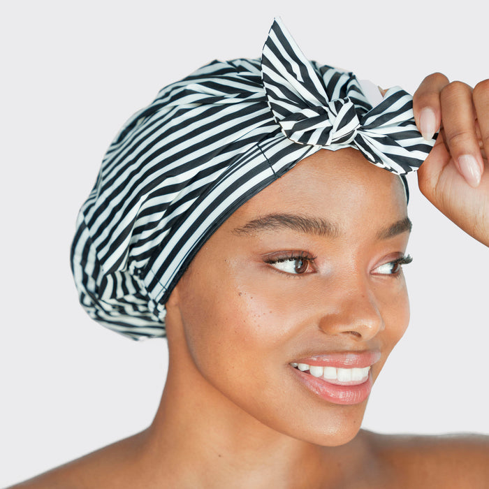 A woman, exuding effortless elegance, smiles while wearing the chic Kitsch Recycled Polyester Luxe Shower Cap in black and white stripes with a bow on top. She looks to the side against a plain background.