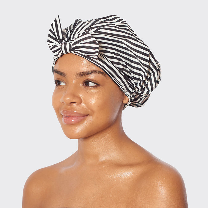A person smiles, displaying the Kitsch Recycled Polyester Luxe Shower Cap, featuring a chic black and white striped design with a bow. The background is light gray.