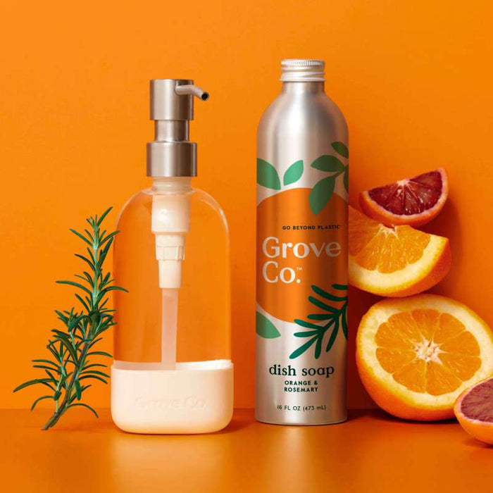 A Refillable Dish Soap Dispenser and a Grove Co. metal bottle with an orange rosemary design stand against an orange backdrop, accented by sliced oranges and rosemary sprigs, promoting sustainability and reducing plastic waste.