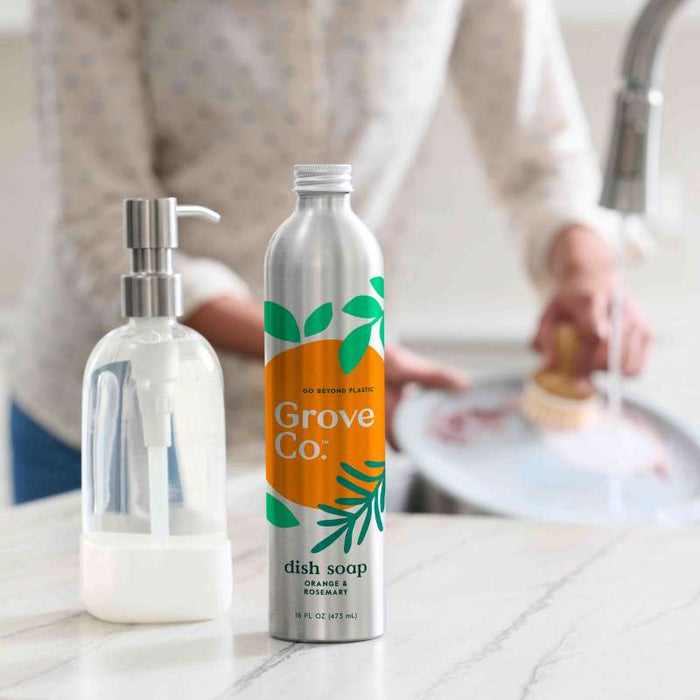 A Grove Co. Refillable Dish Soap Dispenser with orange and rosemary dish soap sits on a kitchen counter as a hand scrubs a pan, promoting sustainability and reducing plastic waste effortlessly.