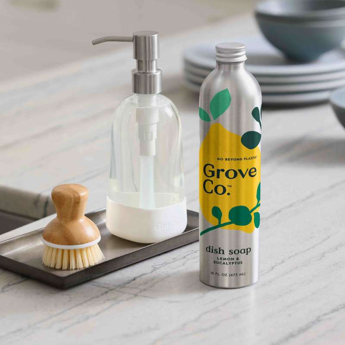 A Grove Co. lemon eucalyptus dish soap bottle, ideal for a sustainable home, sits on the counter in its green and yellow design. Next to it is a Grove Co. refillable dish soap dispenser with a wooden scrub brush on a rectangular tray, all aimed at reducing plastic waste.