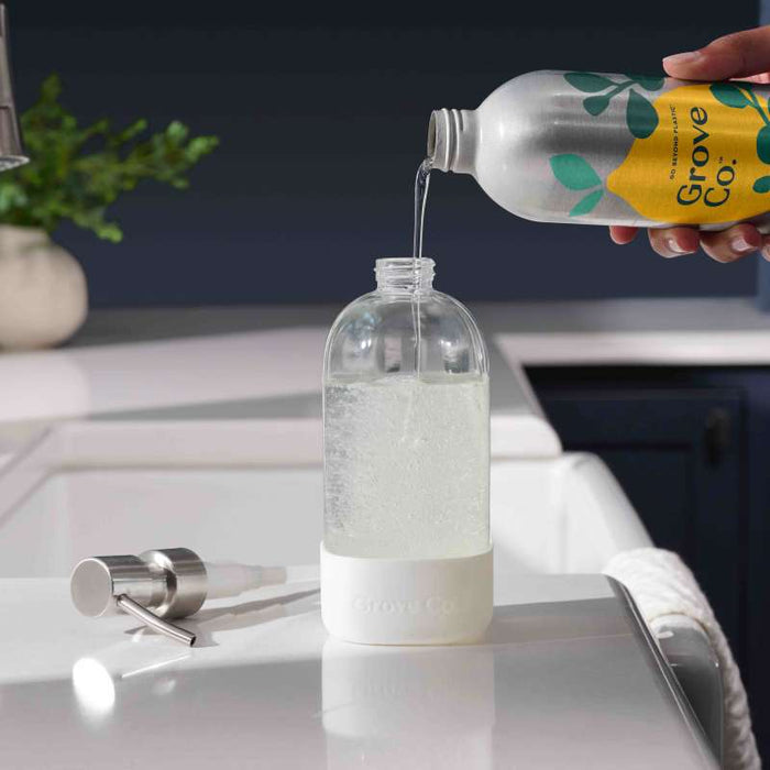 Someone is transferring liquid from a Grove Co. bottle into a Refillable Dish Soap Dispenser on the kitchen counter. A metal pump sits nearby, symbolizing a commitment to reducing plastic waste. The scene includes a sink against a dark wall, highlighting an eco-friendly home environment.