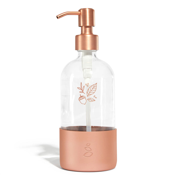 The Grove Co. refillable dish soap dispenser, adorned with a leaf and acorn illustration, features a clear glass body and a rose gold pump and base, combining elegance with eco-friendliness for a sustainable home.