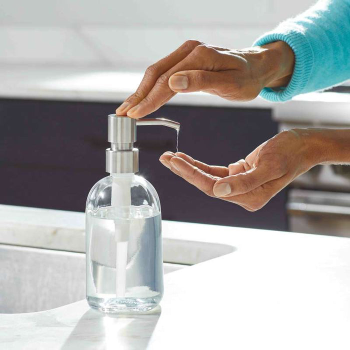 A person in a light blue sleeve uses the Grove Co. Refillable Gel Hand Soap Dispenser, featuring a metallic pump and clear glass container. This eco-friendly dispenser sits elegantly on a white kitchen countertop.