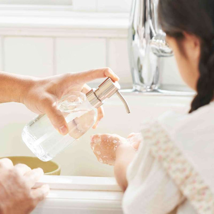 An adult assists a child in washing hands at a sink using the Grove Co. Refillable Gel Hand Soap Dispenser. The child, in a white shirt with floral trim, watches as the adult presses the pump. The eco-friendly scene is complete with a faucet and blurred white tile wall in the background.