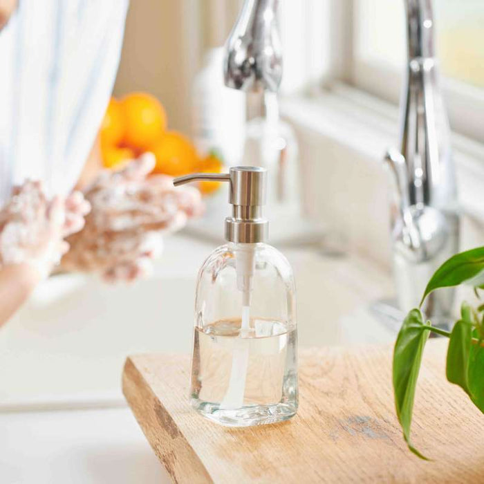 A Grove Co. Refillable Gel Hand Soap Dispenser sits on a wooden counter near a kitchen sink, where someone is washing their hands. In the background, oranges and a green plant enhance the sustainable scene.
