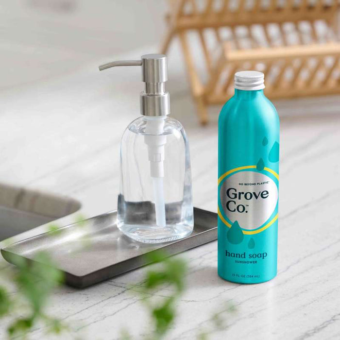 A Grove Co. Refillable Gel Hand Soap Dispenser pairs with a Blue Grove Co. hand soap bottle labeled Sunshine on the kitchen counter, framed by a wooden dish rack and vibrant green plant leaves, enhancing this eco-friendly scene.