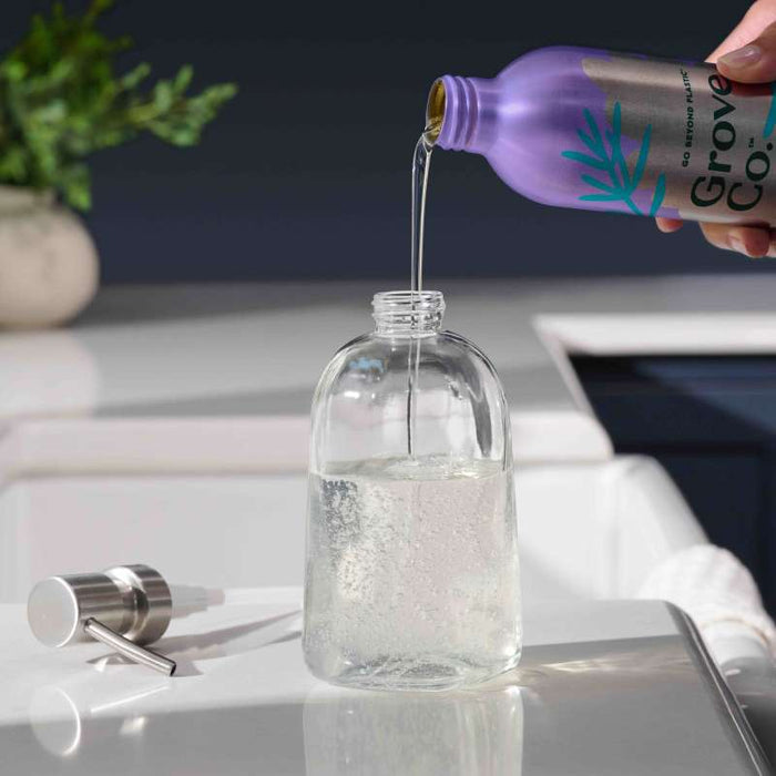 A person pours liquid from a purple bottle into Grove Co.s Refillable Gel Hand Soap Dispenser on the kitchen counter, with the dispenser pump beside it and a plant blurred in the background to highlight the scenes sustainable vibe.