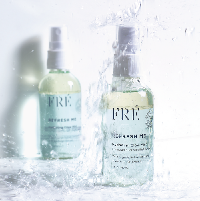 Two clear bottles of FRÉ Skincares REFRESH ME Toning Glow Mist, containing hyaluronic acid, with white spray tops and light green liquid inside, are splashed with water. This enhances the refreshing vibe, ideal for any skincare routine.