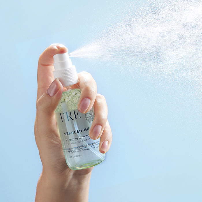 A hand holds a clear bottle with a white nozzle, spraying a fine mist of REFRESH ME Toning Glow Mist by FRÉ Skincare against a light blue background. The label reads Refresh Me, containing green liquid ideal for any skincare routine.