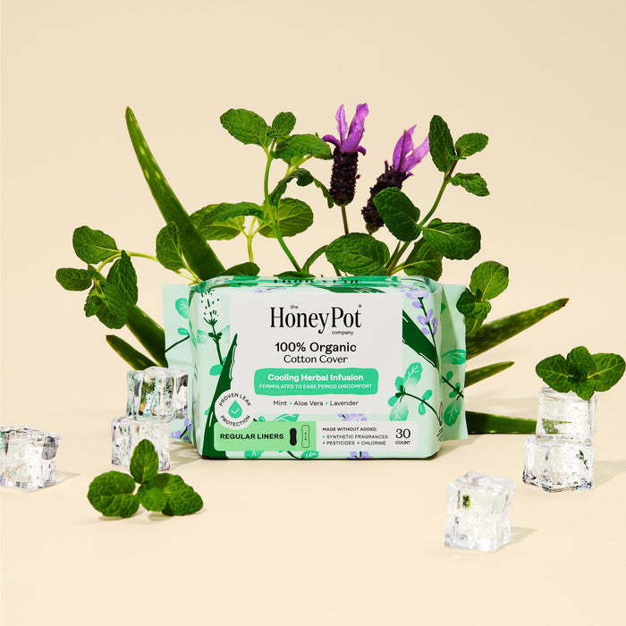 A box of The Honeypot Company 100% Organic Cotton Cover Herbal-Infused Pantiliners is shown on a beige background, surrounded by aloe leaves, mint sprigs, lavender, and ice cubes.