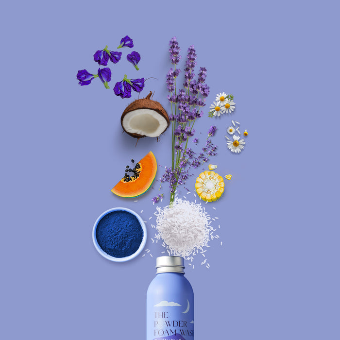 A blue bottle labeled Relaxing Night Body Foam Wash by The Powder Shampoo sits elegantly on a muted purple background, surrounded by ingredients: vibrant butterfly pea flower extract, purple flowers, coconut halves, lavender sprigs, chamomile, a papaya slice, honeycomb, blue powder, and shredded coconut.