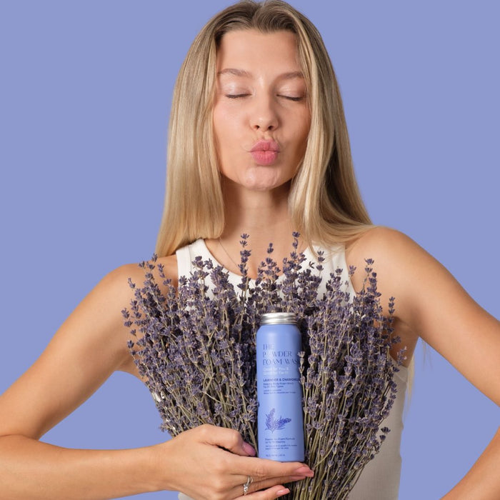 A person with long blonde hair in a white top holds a bouquet of lavender and a lavender can labeled Relaxing Night Body Foam Wash by The Powder Shampoo. The light purple backdrop enhances the serene scene as they pucker their lips, savoring the calming scent of lavender essential oils.