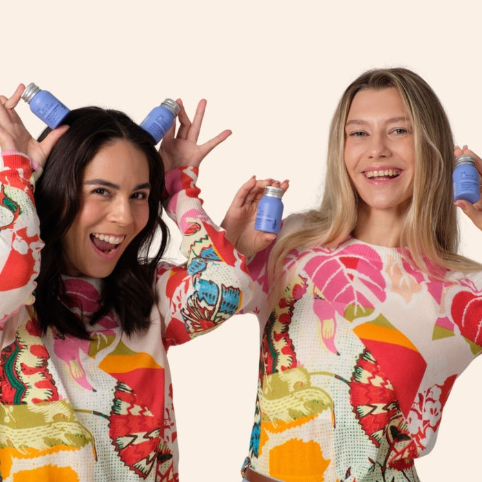 Two women smile brightly, holding recyclable aluminum bottles of The Powder Shampoos Relaxing Night Body Foam Wash near their heads, wearing colorful floral-patterned sweaters against a plain light background.