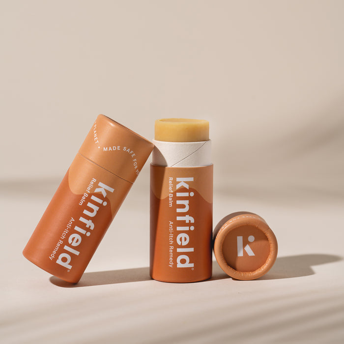 Two orange Kinfield Relief Balm tubes appear, one standing with its cap off to reveal the balm and the other lying flat. The packaging highlights its anti-itch formula featuring cajeput and colloidal oatmeal against a soothing, softly-focused background.