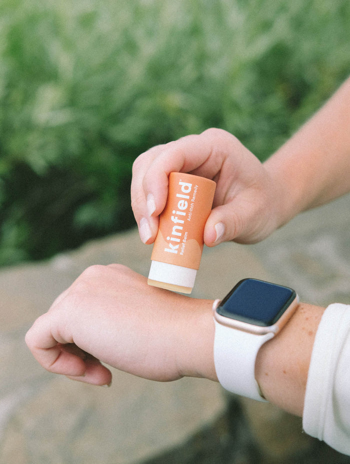 Someone applies Kinfield Relief Balm to their wrist outdoors. They wear a white smartwatch on the left wrist and a light long-sleeve shirt. In the background, blurred greenery mingles with a stone surface, setting a serene scene.