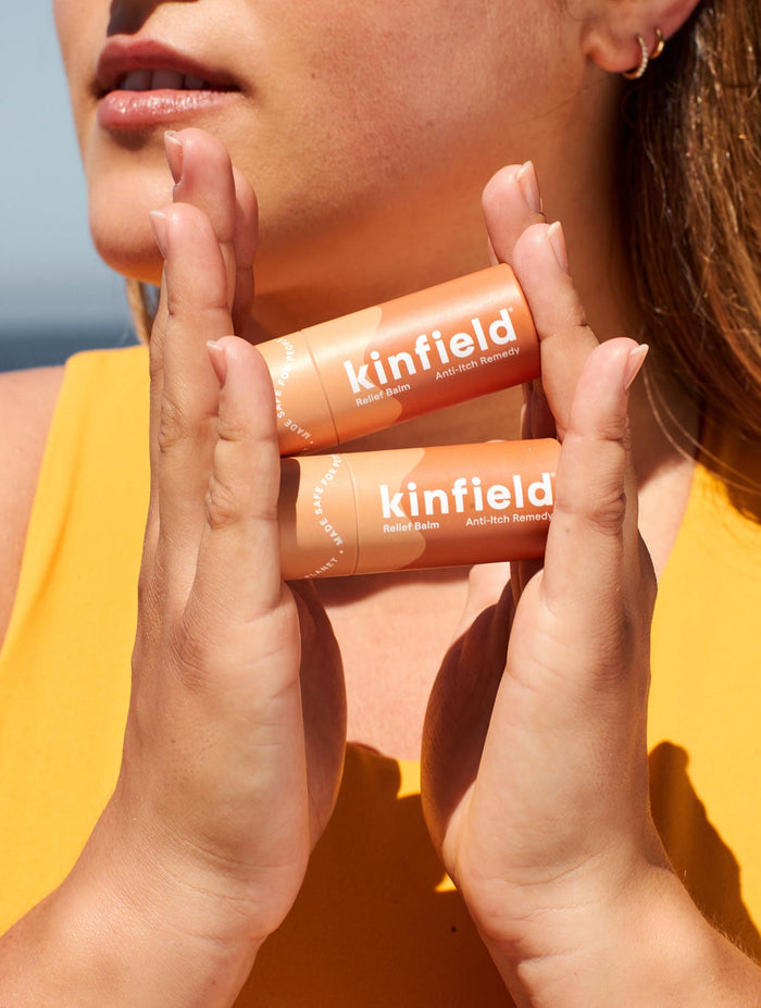 A person holds two orange Kinfield Relief Balm tubes, labeled Anti-Itch Remedy and infused with cajeput, against their chest. Theyre wearing a yellow top under a clear blue sky.