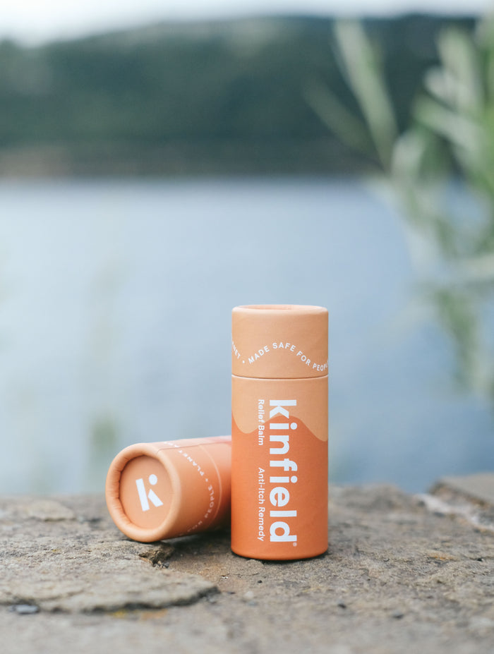Two Kinfield Relief Balm tubes are placed on a stone surface by a calm water body, one upright and the other lying down. The background has a blurred view of glistening water and verdant greenery, creating a serene atmosphere.