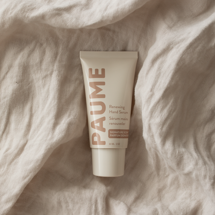 A tube of PAUMEs Renewing Hand Serum, rich in Vitamin C and Hyaluronic Acid, lies on textured beige fabric. The light-colored packaging displays the product name and description in soft, earthy tones.
