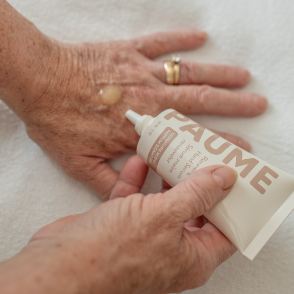 A persons hand applies a small amount of PAUME Renewing Hand Serum onto the back of their other hand, highlighting a wedding ring on a white surface as its hyaluronic acid-infused formula works wonders.