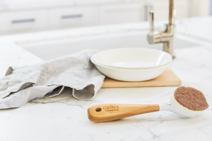 A Grove Co. Replaceable Head Walnut Scrubber Dish Brush with a wooden handle sits on a marble countertop. A white dish is on a wooden board nearby, next to a crumpled gray cloth by the stainless steel sink faucet, ideal for non-abrasive cleaning.