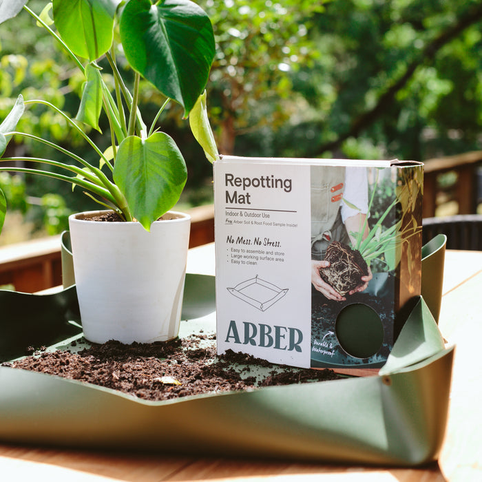 An eco-friendly green Arber Repotting Mat holds a white potted plant with spilled soil, while a box labeled Arber Repotting Mat features hands tending to plants, set against a wooden deck and lush greenery for an organic touch.