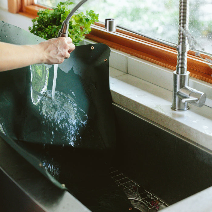 An individual uses a flexible faucet sprayer to rinse a large, Arber Repotting Mat in the kitchen sink. The eco-friendly setup features a green plant on the windowsill under the wooden-framed window, with water splashing over the black mat and into the sink.