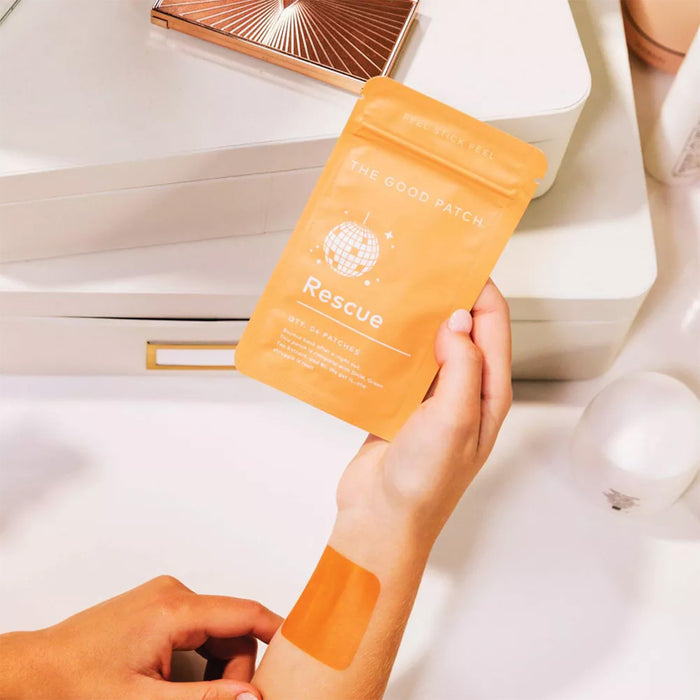 A person applies an orange Rescue Patch by The Good Patch on their arm, next to its matching packet. A white surface with a makeup compact and toiletries depicts the revitalizing touch of vitamin B1 in each hangover patch.