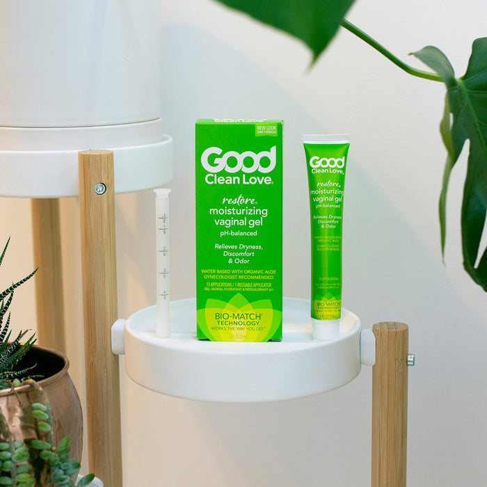 The image shows a green box and matching tube of Good Clean Loves Restore pH-Balancing & Moisturizing Vaginal Gel on a white circular shelf, surrounded by lush plants, emphasizing its pH-balanced formula for vaginal health.