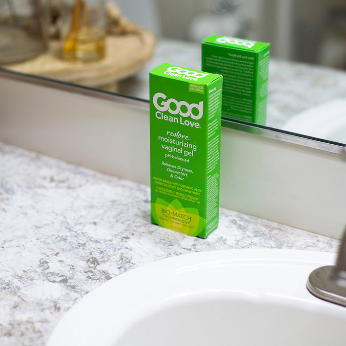 A box of Good Clean Love Restore pH-Balancing & Moisturizing Vaginal Gel sits on a bathroom countertop near the sink. Its green packaging with white text is reflected in the mirror, while a blurred background highlights a tray of toiletries, underlining its focus on vaginal health.