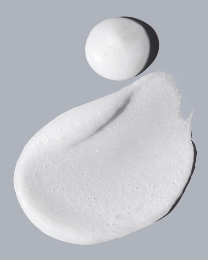 On a soft gray background, a smooth swirl and round dollop of white cream showcase the texture and consistency of Acures Resurfacing Glycolic & Unicorn Root Cleanser.