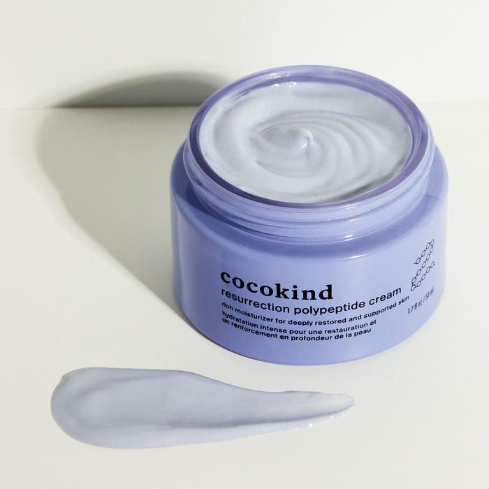 An open jar of Cocokinds Resurrection Polypeptide Cream reveals a smooth, lavender-colored cream promising deep hydration. A small dab rests on the white surface beyond the jar, casting a light shadow as it awaits to tackle fine lines and wrinkles.