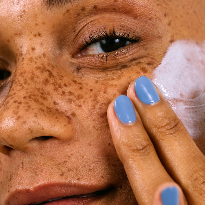 Close-up of a freckled person applying Cocokinds Resurrection Polypeptide Cream to their cheek, enhancing hydration. Their blue-painted nails gently glide across the skin, visible alongside long lashes. The complexion appears smooth and deeply moisturized.